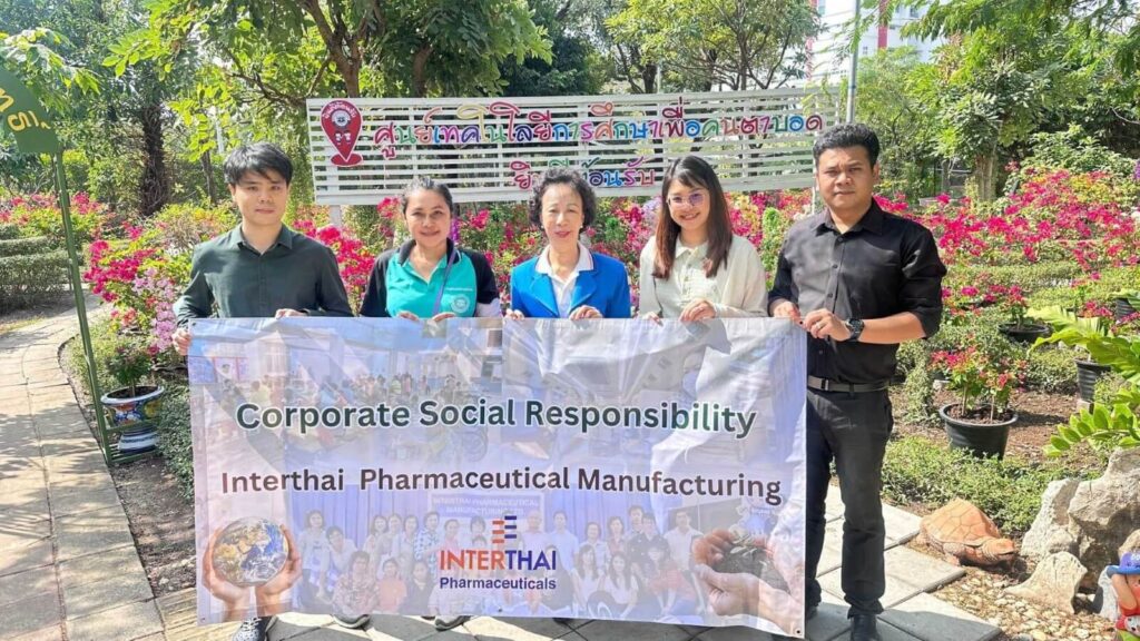 Five people stand holding a banner that reads "Corporate Social Responsibility Interthai Pharmaceutical Manufacturing" with images of the earth and people.